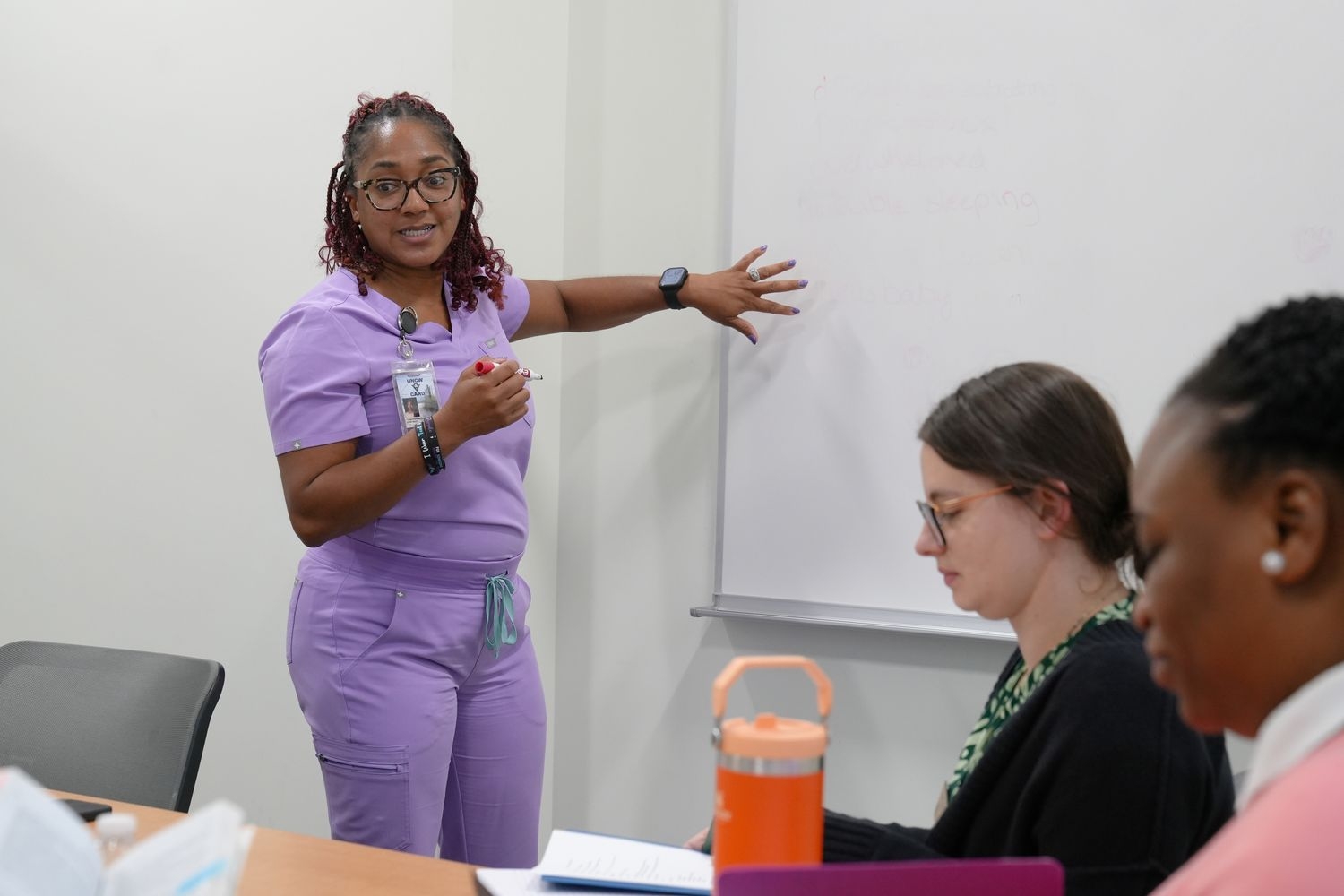 A DNP student in scrubs stands in front of a blank whiteboard with a marker, ready to write her and two fellow DNP students notes during their intensive practice.