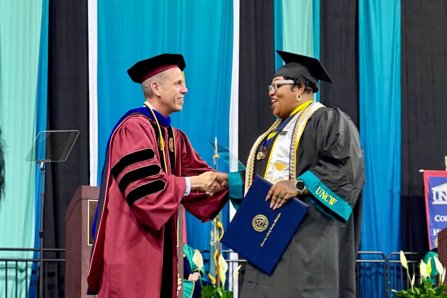 An HS2SW in their cap and gown (right) shakes hands with College of Health and Human Services Dean Jack Watson (left) on stage at commencement.