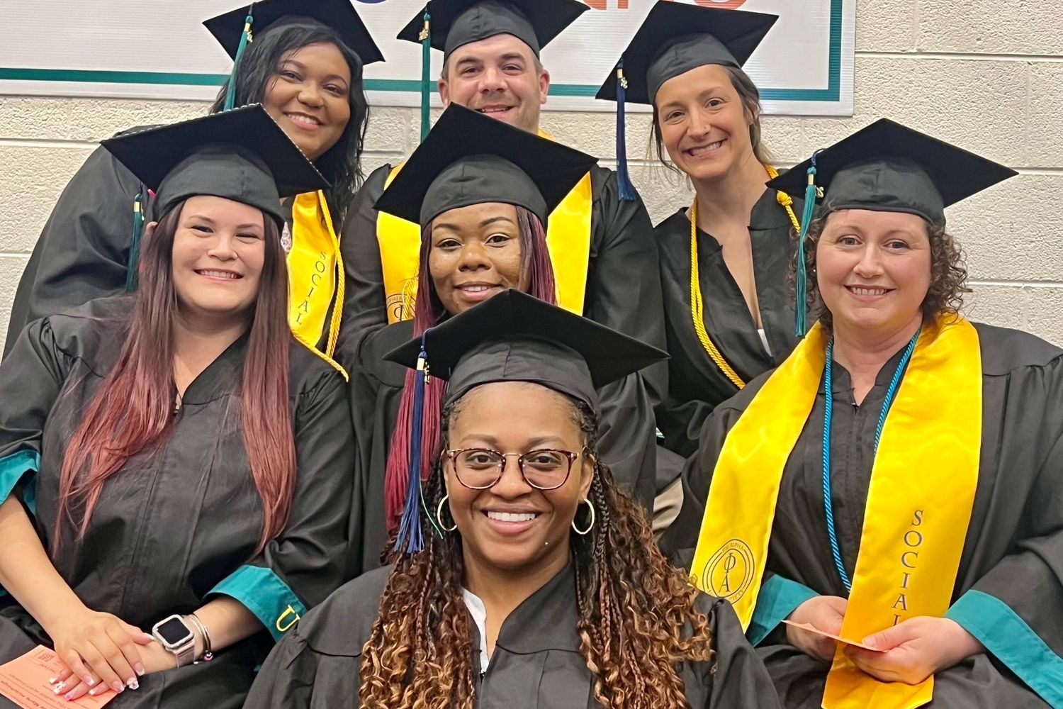 A group of seven HS2SW social work graduates in their caps and gowns pose for a photo on the steps inside Trask Coliseum during commencement.