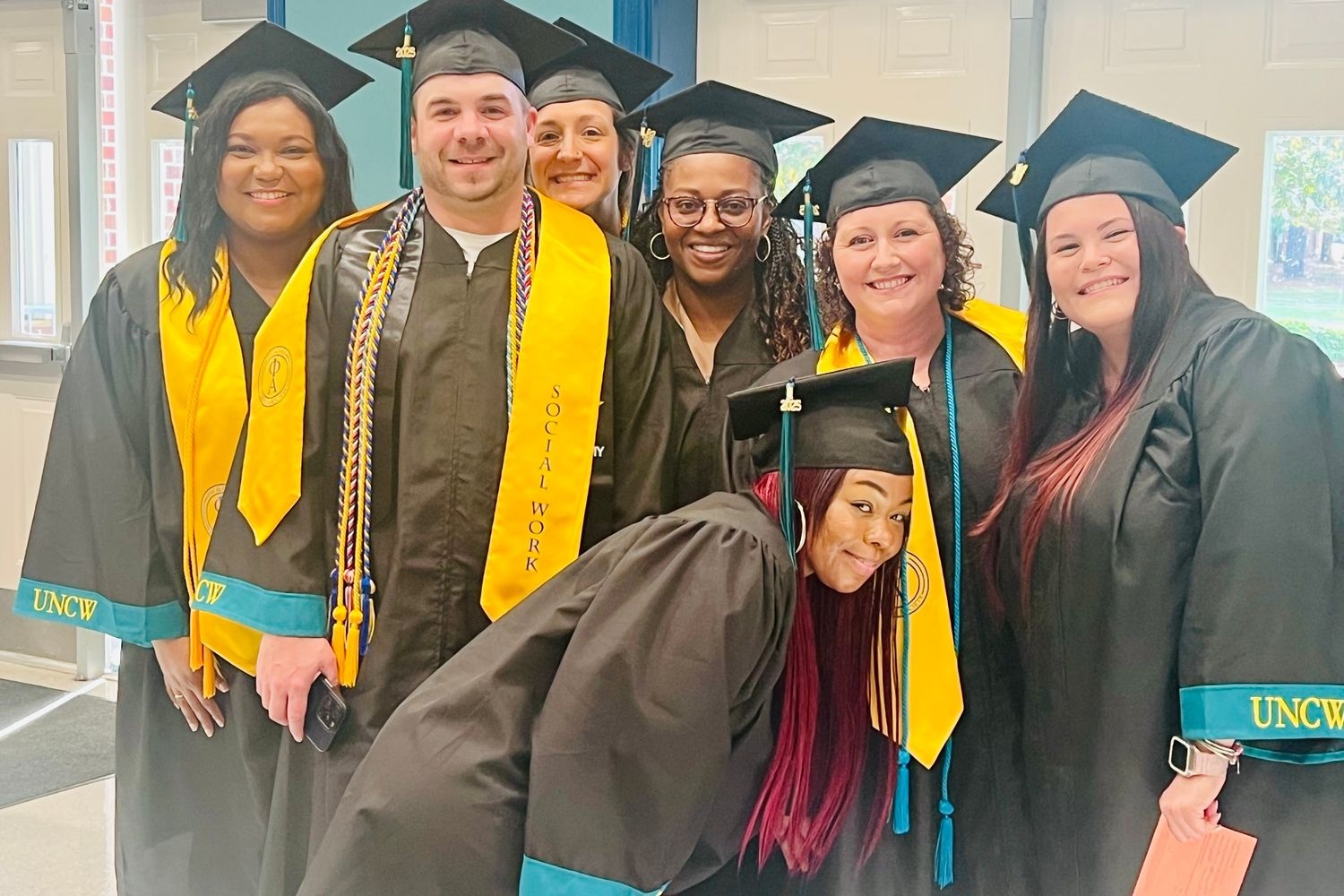 A group of seven HS2SW social work graduates in their caps and gowns pose for a photo inside Trask Coliseum during commencement.