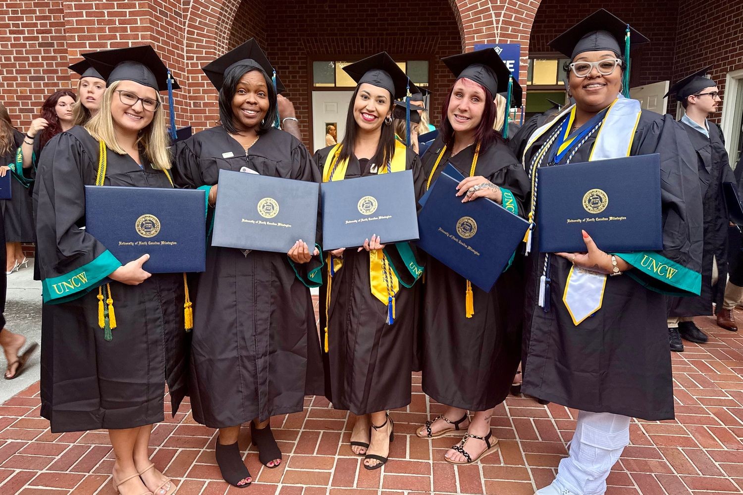 A group of five HS2SW social work graduates in their caps and gowns pose for a photo outside Trask Coliseum after commencement. Each one is holding their diploma holder.