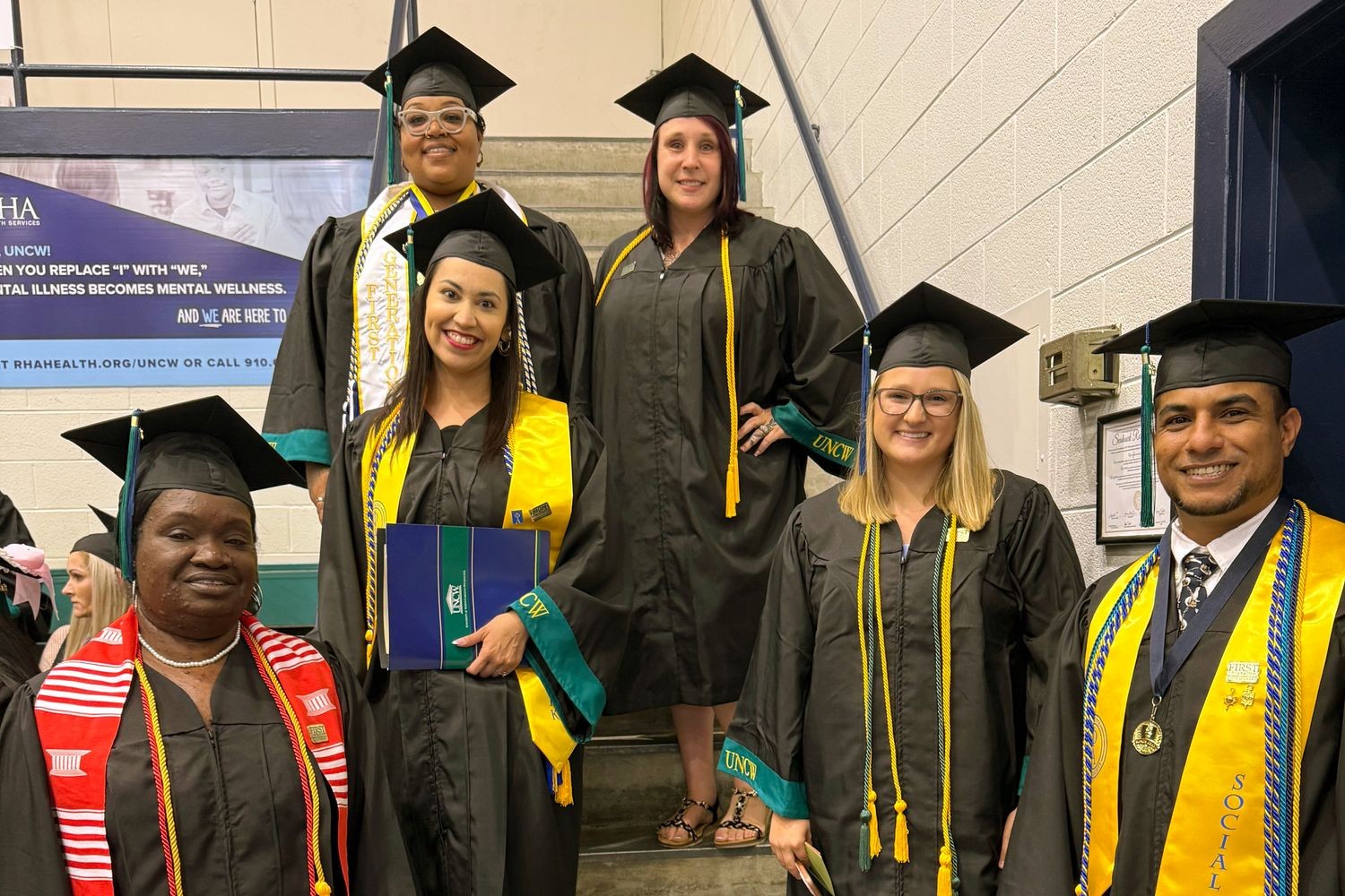 A group of six HS2SW social work graduates in their caps and gowns pose for a photo on the steps inside Trask Coliseum during commencement.